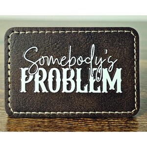 May include: A brown leather patch with white stitching around the edges. The text "Somebody's PROBLEM" is printed in white on the patch.