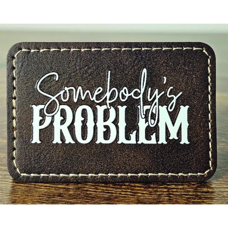 May include: A brown leather patch with white stitching around the edges. The text "Somebody's PROBLEM" is printed in white on the patch.