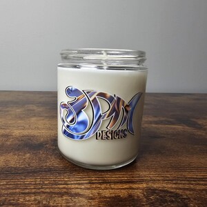 May include: A white candle in a clear glass jar with a blue and gold swirl design and the text "3PM Designs".