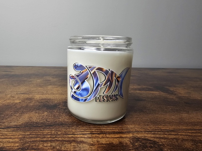 May include: A white candle in a clear glass jar with a blue and gold swirl design and the text "3PM Designs".