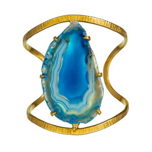 May include: A gold-toned cuff bracelet featuring a large, teardrop-shaped blue agate stone. The stone has concentric bands of blue, white, and tan. The bracelet has a textured band and curved supports that hold the stone in place.