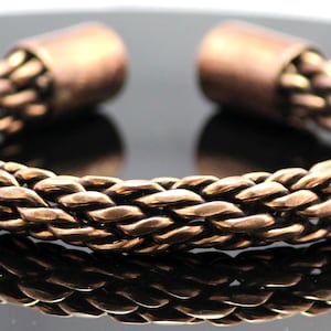 Peut inclure: Un bracelet manchette de couleur cuivre avec un motif tressé. Le bracelet a une forme arrondie avec deux embouts solides. La section tressée est composée de plusieurs brins tissés ensemble. Le bracelet est présenté sur une surface réfléchissante.
