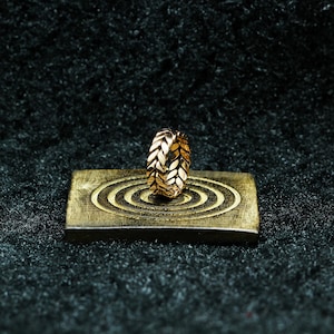 Peut inclure: Une bague dorée présentant un motif répétitif de feuilles ou d'épis de blé sur tout l'anneau. La bague est placée verticalement sur une base rectangulaire de couleur métallique, ornée d'une série de rainures circulaires concentriques.