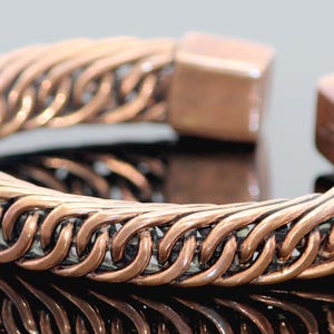 Pode incluir: Uma pulseira de punho cor de cobre com um design de corrente trançada. A pulseira tem um fecho quadrado em cada extremidade. O metal reflete a luz, criando uma aparência brilhante. Esta joia é um acessório elegante.