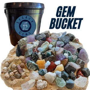 May include: A black plastic bucket with a blue circular label reading Legacy Gems Est. 2023, accompanied by a large pile of assorted raw gemstones, minerals, and small seashells scattered across a tan sandy base, with the text GEM BUCKET nearby.