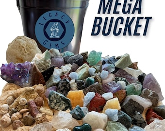 Op de afbeelding: Een zwarte plastic emmer met het label Legacy Gems Mega Bucket gevuld met een verscheidenheid aan ruwe mineralen, kristallen en schelpen. De collectie bevat amethistclusters, kwarts, groene fluoriet en diverse stenen verspreid op zand.