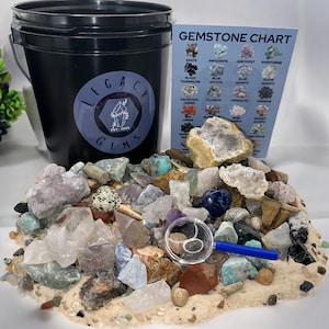 May include: A black bucket filled with a variety of colorful gemstones and a magnifying glass. The bucket has a white sticker with the text "LEGACY GEMS EST. 2023". A chart with the text "GEMSTONE CHART" is visible in the background.