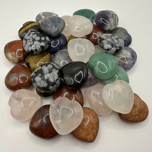 May include: A collection of heart-shaped polished stones in various colors and patterns, including pink, green, black, brown, and white. The stones are arranged in a pile on a white surface.