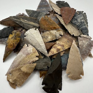 May include: A collection of approximately 30 arrowheads made of various types of stone. The arrowheads are in shades of brown, gray, and black. Some of the arrowheads have a mottled or banded pattern.