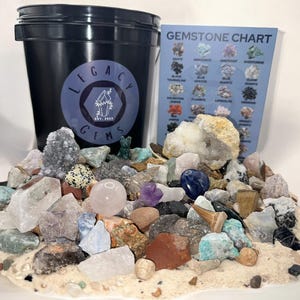 May include: A black bucket filled with a variety of colorful gemstones and crystals. The bucket has a sticker with the text "Legacy Gems Est. 2023". A gemstone chart is visible in the background.