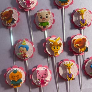 Pu&ograve; includere: Topper per cupcake con vari personaggi dei cartoni animati. Ogni topper ha una base a forma di fiore rosa con un centro a pois e uno stelo trasparente. I personaggi includono un orso, un maiale, una giraffa e altri.