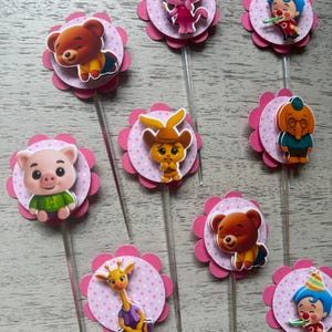 Pu&ograve; includere: Una collezione di decorazioni per cupcake colorate con vari personaggi dei cartoni animati. Ogni decorazione presenta un'immagine del personaggio su un cerchio rosa a pois, circondato da un fiore rosa smerlato. Sono attaccati bastoncini di plastica trasparente.