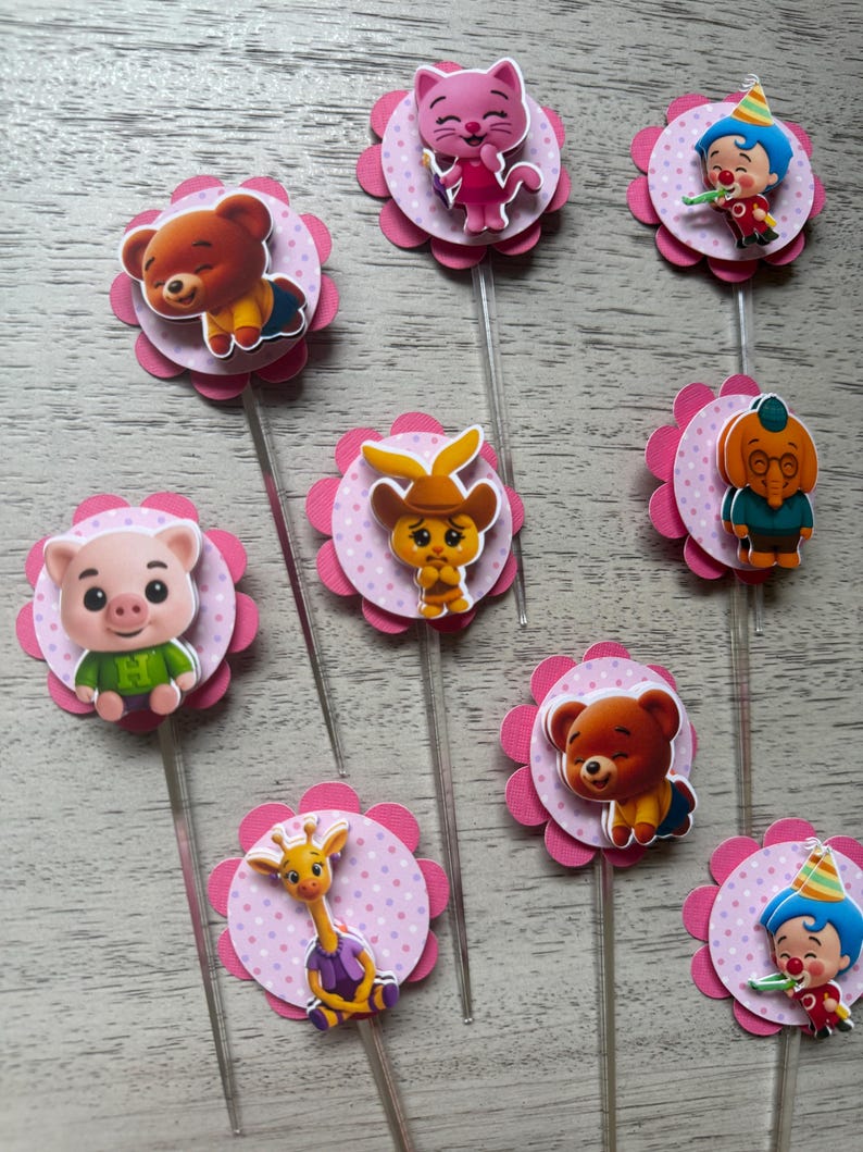 Pu&ograve; includere: Una collezione di decorazioni per cupcake colorate con vari personaggi dei cartoni animati. Ogni decorazione presenta un'immagine del personaggio su un cerchio rosa a pois, circondato da un fiore rosa smerlato. Sono attaccati bastoncini di plastica trasparente.
