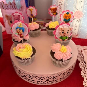 Pu&ograve; includere: Una collezione di cupcakes con glassa rosa e gialla, ognuno sormontato da una decorazione a tema personaggio. I cupcakes sono disposti su un alzatina bianca e decorativa, su sfondo rosso.