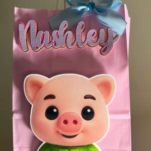 Puede incluir: Bolsa de regalo rosa con una ilustraci&oacute;n de un cerdo de dibujos animados con una camisa verde y pantalones cortos morados. La bolsa tiene el nombre "Nashley" en cursiva, un lazo azul claro y asas blancas.