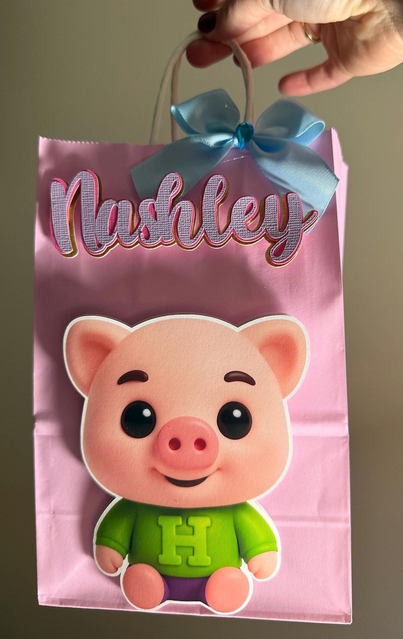 Puede incluir: Bolsa de regalo rosa con una ilustraci&oacute;n de un cerdo de dibujos animados con una camisa verde y pantalones cortos morados. La bolsa tiene el nombre "Nashley" en cursiva, un lazo azul claro y asas blancas.
