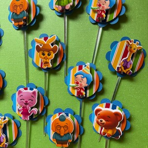May include: A collection of colorful cupcake toppers featuring various cartoon characters. Each topper has a character attached to a striped circle with a blue scalloped edge, mounted on a clear plastic stick. The characters include a pig, clown, and giraffe.