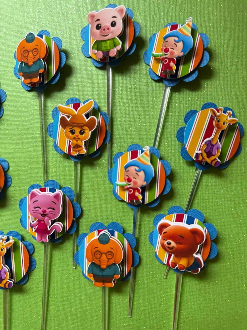 May include: A collection of colorful cupcake toppers featuring various cartoon characters. Each topper has a character attached to a striped circle with a blue scalloped edge, mounted on a clear plastic stick. The characters include a pig, clown, and giraffe.