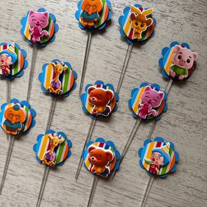 May include: A collection of colorful cupcake toppers featuring various cartoon animal characters. Each topper has a blue scalloped edge, a striped background, and a clear plastic stick. The characters include a cat, bear, giraffe, and pig.