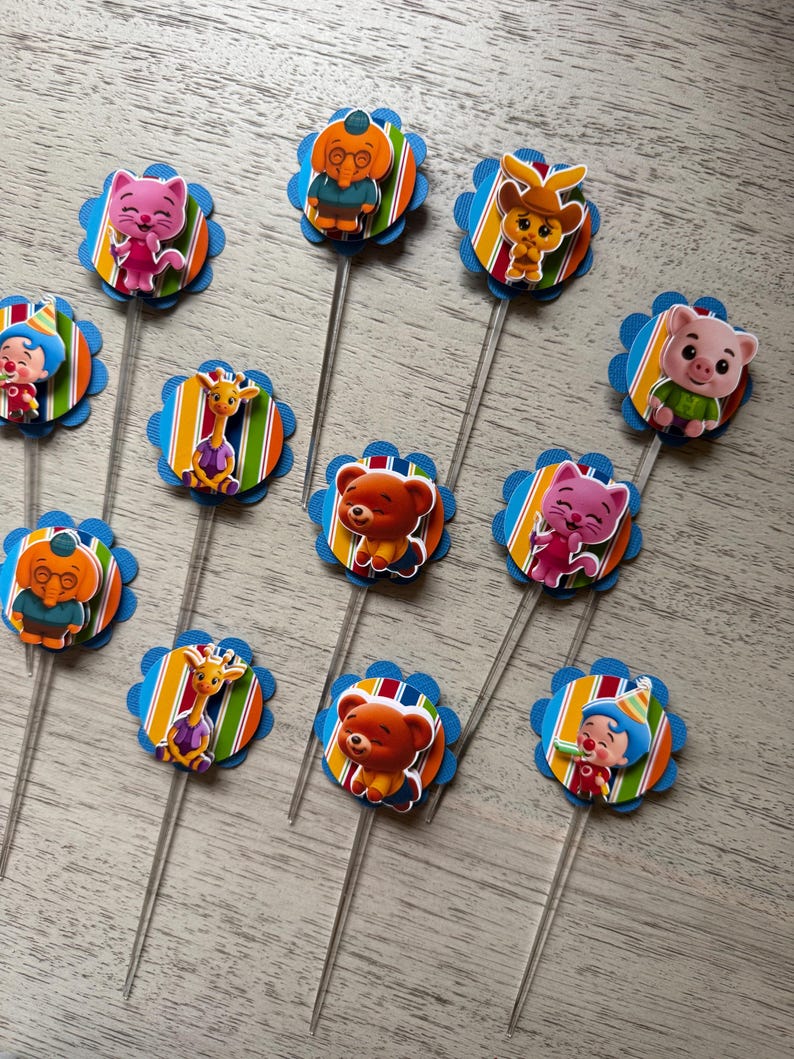 May include: A collection of colorful cupcake toppers featuring various cartoon animal characters. Each topper has a blue scalloped edge, a striped background, and a clear plastic stick. The characters include a cat, bear, giraffe, and pig.
