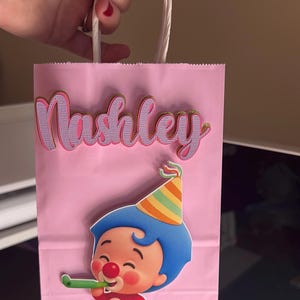 Puede incluir: Bolsa de fiesta rosa con asas de cuerda blanca. La bolsa presenta el nombre "Nashley" en letras rosas y doradas. Un personaje de dibujos animados con pelo azul y un gorro de fiesta tambi&eacute;n est&aacute; en la bolsa.