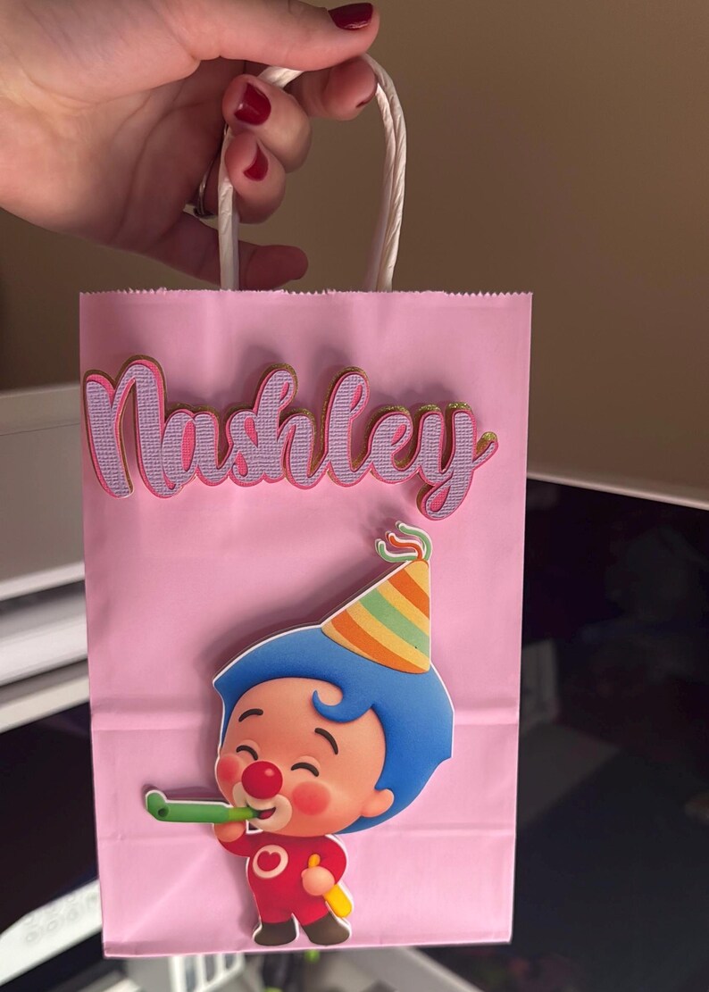Puede incluir: Bolsa de fiesta rosa con asas de cuerda blanca. La bolsa presenta el nombre "Nashley" en letras rosas y doradas. Un personaje de dibujos animados con pelo azul y un gorro de fiesta tambi&eacute;n est&aacute; en la bolsa.