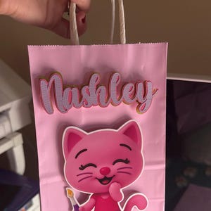 Puede incluir: Bolsa de regalo rosa con el nombre "Nashley" en letra cursiva. Un gato de dibujos animados rosa con un pincel est&aacute; en la parte delantera. La bolsa tiene asas de papel blanco.