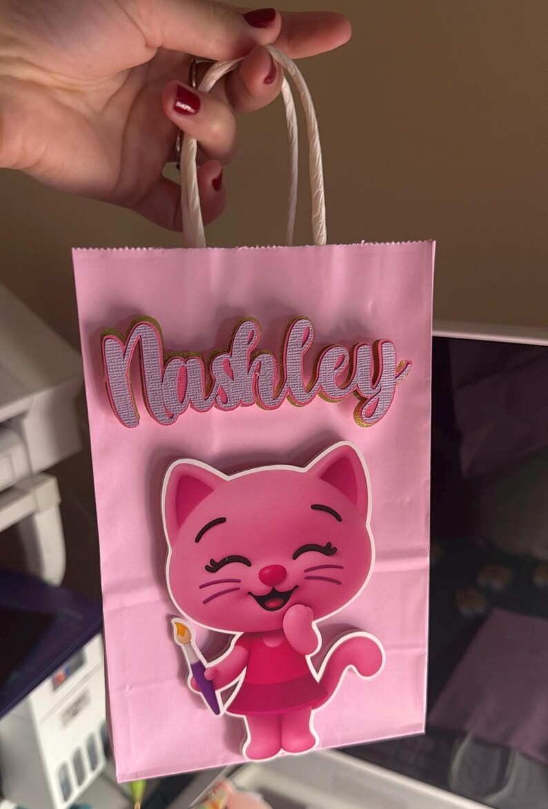 Puede incluir: Bolsa de regalo rosa con el nombre "Nashley" en letra cursiva. Un gato de dibujos animados rosa con un pincel est&aacute; en la parte delantera. La bolsa tiene asas de papel blanco.