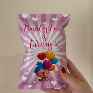 May include: Pink party favor bag with a cartoon clown holding heart-shaped balloons. The bag has a radial design with white hearts and the text "Nashley Eun is Turning 2".