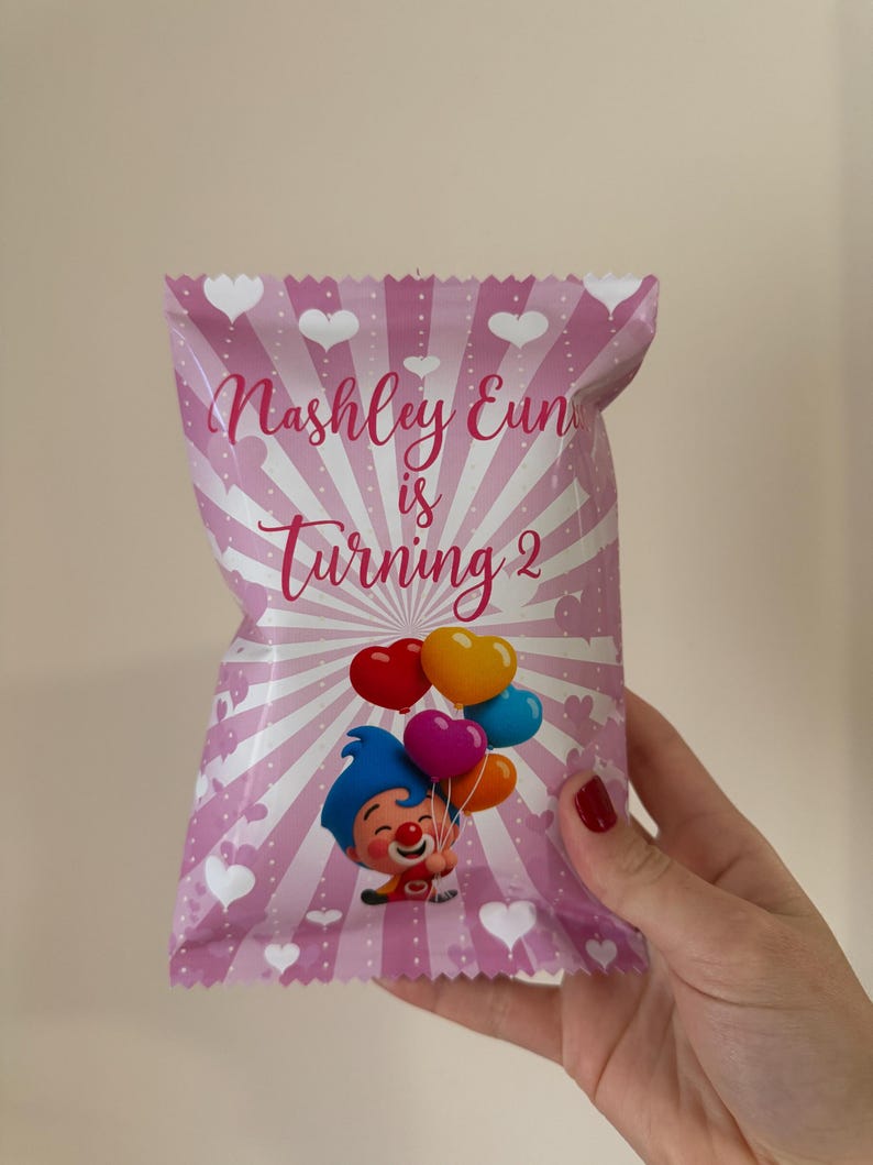 May include: Pink party favor bag with a cartoon clown holding heart-shaped balloons. The bag has a radial design with white hearts and the text "Nashley Eun is Turning 2".