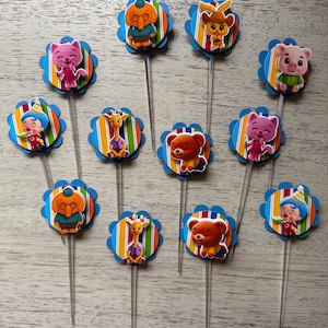 May include: A collection of colorful cupcake toppers featuring various cartoon characters. Each topper has a blue scalloped edge, a striped background, and a clear plastic stick. The characters include a cat, bear, giraffe, and pig.