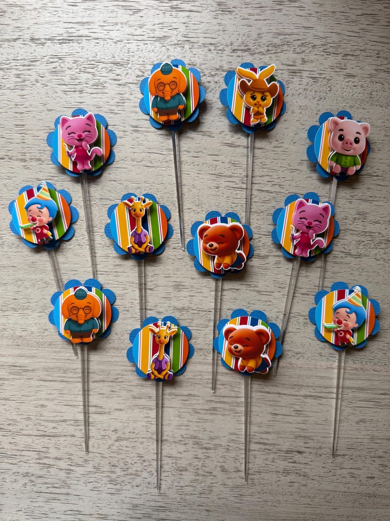 May include: A collection of colorful cupcake toppers featuring various cartoon characters. Each topper has a blue scalloped edge, a striped background, and a clear plastic stick. The characters include a cat, bear, giraffe, and pig.