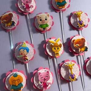 Pu&ograve; includere: Una collezione di decorazioni per cupcake con vari personaggi dei cartoni animati. Ogni decorazione ha un bordo smerlato rosa e un centro rosa chiaro a pois. I personaggi includono un orso, un maiale, un gatto, una giraffa e un clown, tutti attaccati a bastoncini trasparenti.
