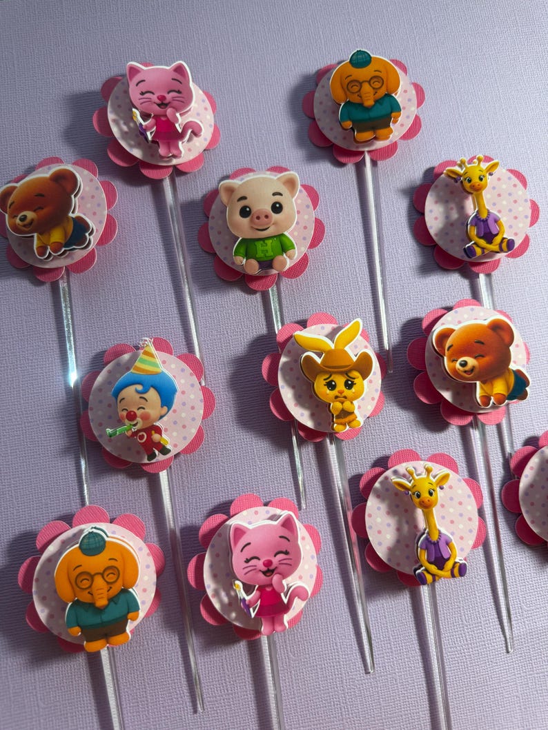 Pu&ograve; includere: Una collezione di decorazioni per cupcake con vari personaggi dei cartoni animati. Ogni decorazione ha un bordo smerlato rosa e un centro rosa chiaro a pois. I personaggi includono un orso, un maiale, un gatto, una giraffa e un clown, tutti attaccati a bastoncini trasparenti.