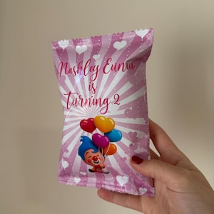 May include: Pink party favor bag with white hearts and radial stripes. Text reads "Nashley Eunice is Turning 2". Features a cartoon clown holding heart-shaped balloons in red, yellow, purple, and blue.
