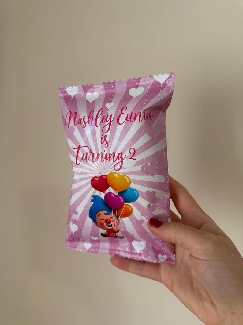 May include: Pink party favor bag with white hearts and radial stripes. Text reads "Nashley Eunice is Turning 2". Features a cartoon clown holding heart-shaped balloons in red, yellow, purple, and blue.
