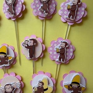 May include: Cupcake toppers with a cartoon monkey design. The toppers feature a pink, scalloped flower shape with a white and yellow patterned background. Some monkeys are holding bananas or wearing yellow hats.