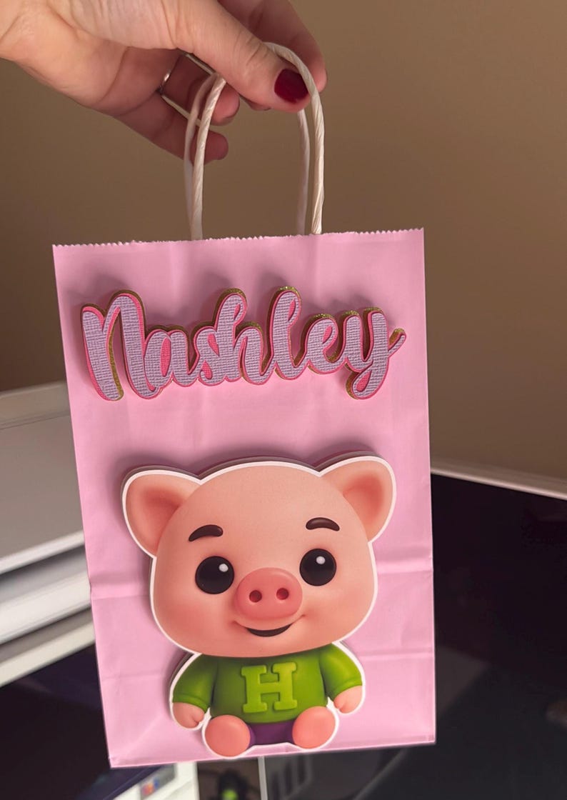 Puede incluir: Bolsa de regalo rosa con el nombre "Nashley" en fuente cursiva con ribete dorado. Un cerdo de dibujos animados con una camisa verde con la letra "H" est&aacute; en la parte delantera de la bolsa. La bolsa tiene asas de cuerda blanca.