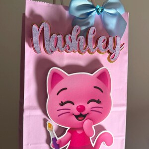 Puede incluir: Bolsa de regalo rosa con el nombre "Nashley" en letras superpuestas. Una ilustraci&oacute;n de gato de dibujos animados est&aacute; en la parte delantera, sosteniendo un pincel. Un lazo azul claro con un centro de joya adorna la parte superior de la bolsa.