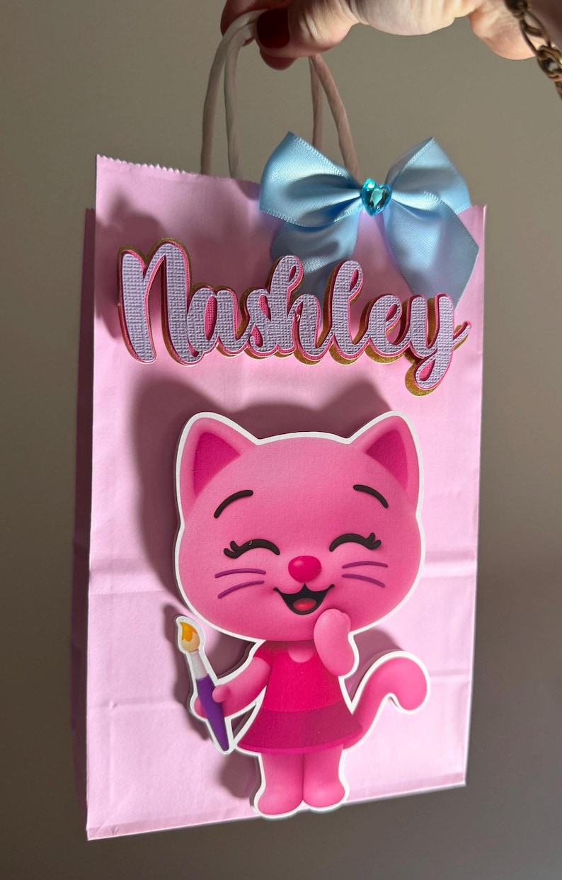 Puede incluir: Bolsa de regalo rosa con el nombre "Nashley" en letras superpuestas. Una ilustraci&oacute;n de gato de dibujos animados est&aacute; en la parte delantera, sosteniendo un pincel. Un lazo azul claro con un centro de joya adorna la parte superior de la bolsa.