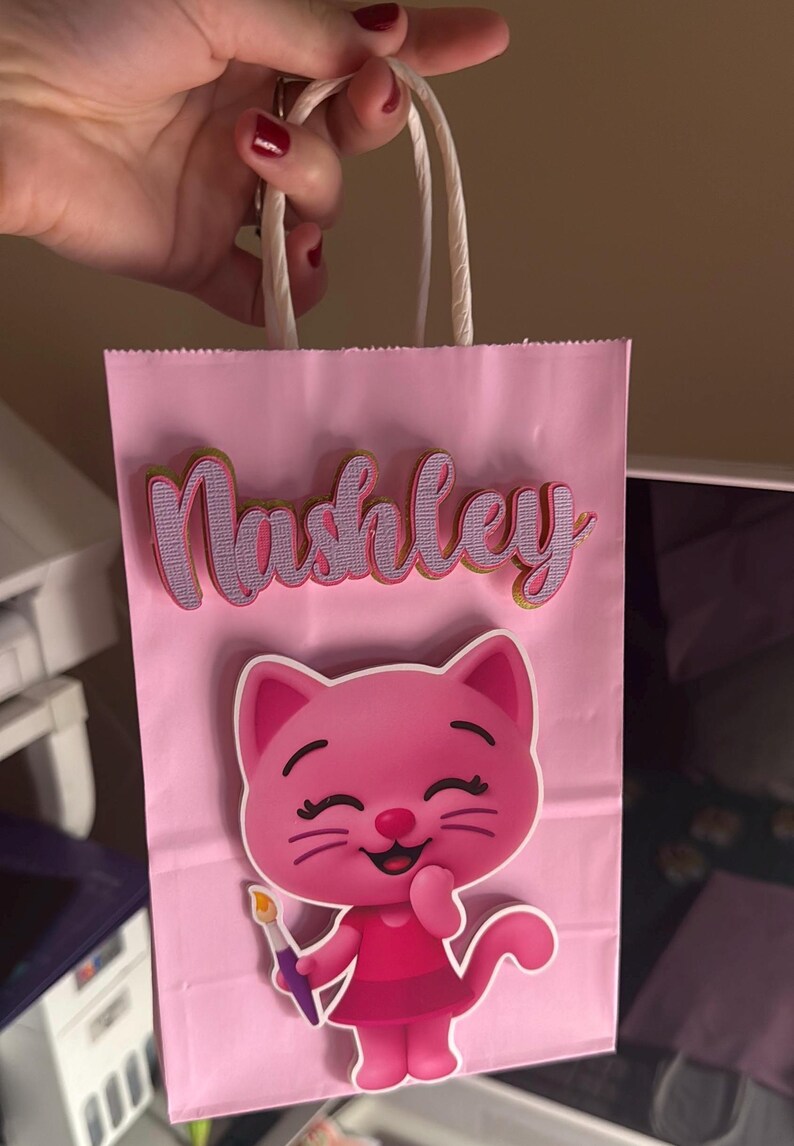 Puede incluir: Bolsa de regalo rosa con el nombre "Nashley" en fuente cursiva. Un personaje de dibujos animados de un gato rosa con un pincel est&aacute; en la parte delantera de la bolsa. La bolsa tiene asas de papel blanco.