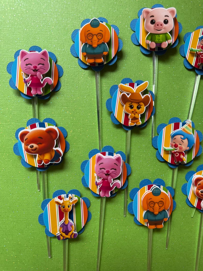 May include: A collection of colorful cupcake toppers featuring various cartoon animal characters. Each topper has a blue scalloped edge, a striped center, and a clear plastic stick. The characters include a pig, cat, bear, elephant, and giraffe.