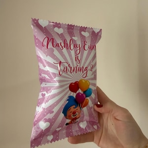 May include: Pink party favor bag with white hearts and stripes, featuring the text "Nashley Eun is Turning 2." A cartoon clown holding colorful balloons is also printed on the bag. The bag is held by a person.
