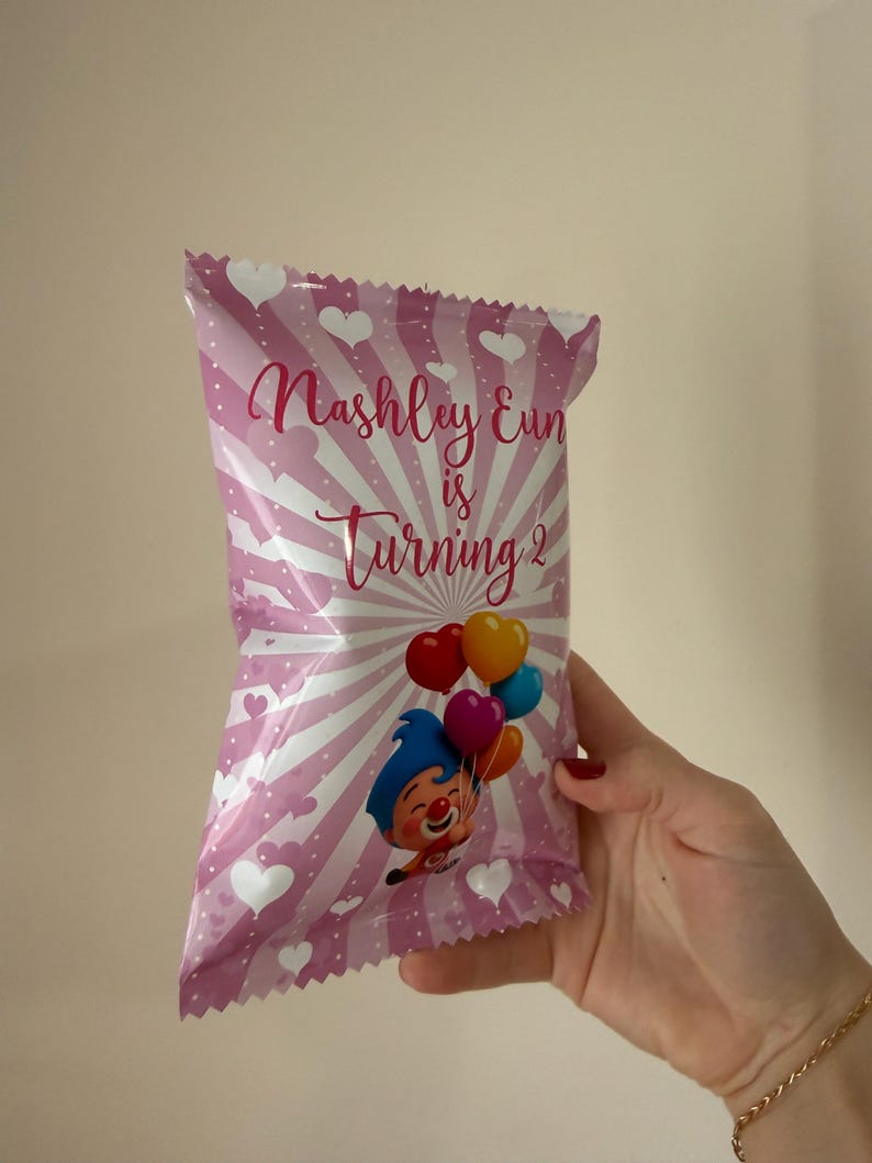 May include: Pink party favor bag with white hearts and stripes, featuring the text "Nashley Eun is Turning 2." A cartoon clown holding colorful balloons is also printed on the bag. The bag is held by a person.