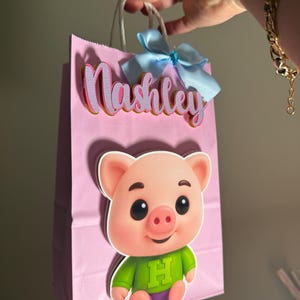 Puede incluir: Bolsa de regalo rosa con una ilustraci&oacute;n de cerdo de dibujos animados y el nombre "Nashley" en letras rosas y doradas. La bolsa tiene un lazo azul claro y asas blancas.