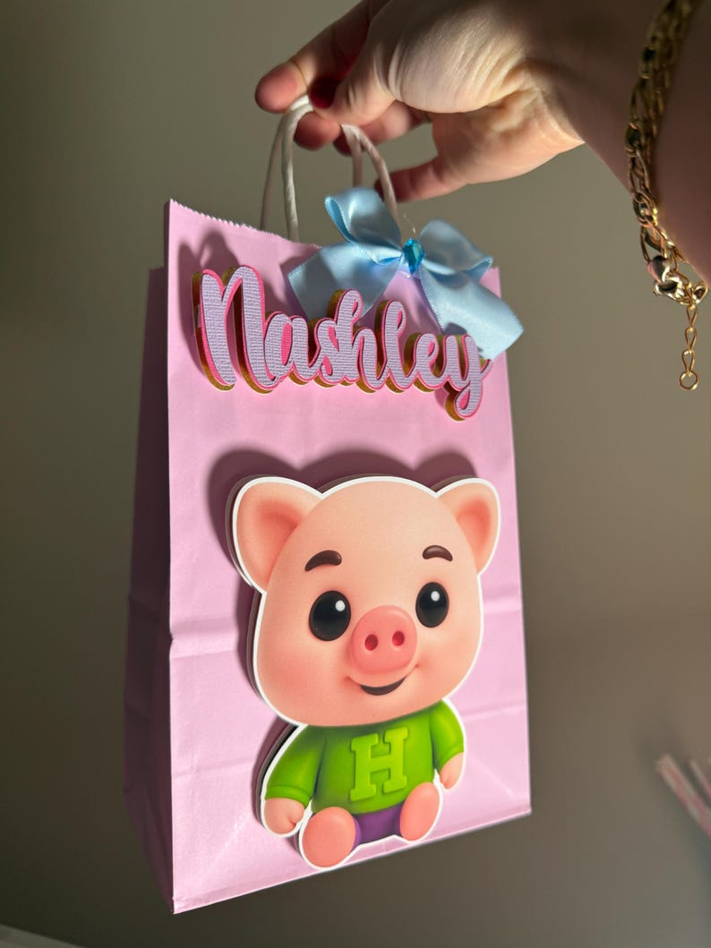 Puede incluir: Bolsa de regalo rosa con una ilustraci&oacute;n de cerdo de dibujos animados y el nombre "Nashley" en letras rosas y doradas. La bolsa tiene un lazo azul claro y asas blancas.