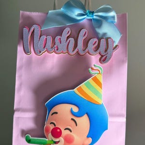 Puede incluir: Bolsa de regalo rosa con el nombre "Nashley" en letras cursivas. Un lazo azul claro adorna la parte superior. La bolsa presenta un payaso de dibujos animados con pelo azul, un gorro de fiesta y un atuendo rojo. El payaso sostiene un silbato verde.