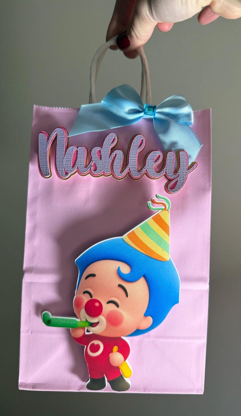 Puede incluir: Bolsa de regalo rosa con el nombre "Nashley" en letras cursivas. Un lazo azul claro adorna la parte superior. La bolsa presenta un payaso de dibujos animados con pelo azul, un gorro de fiesta y un atuendo rojo. El payaso sostiene un silbato verde.