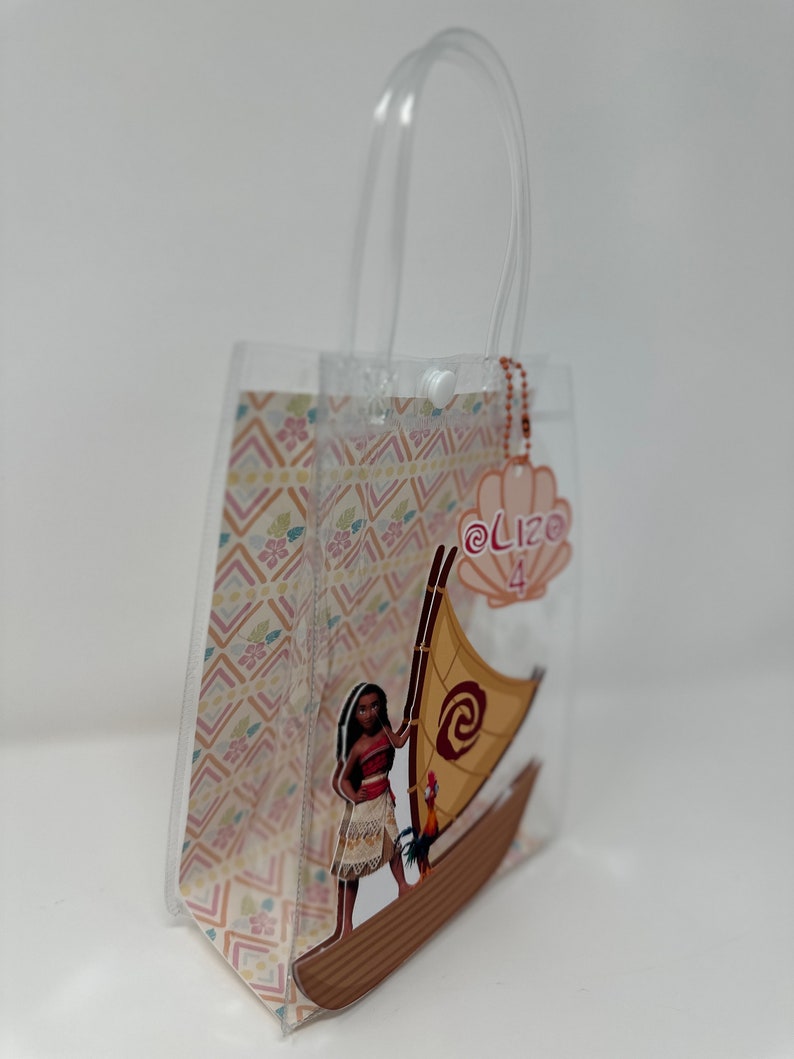 Może przedstawiać: Przezroczysta plastikowa torba z nadrukiem przedstawiającym polinezyjską kobietę z kogutkiem na łodzi. Torba ma biały uchwyt i r&oacute;żową metkę z tekstem "@Lizo 4".