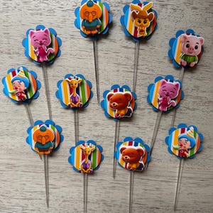May include: A collection of colorful cupcake toppers featuring various cartoon characters. Each topper has a blue scalloped edge, a striped background, and a character image. The toppers are on clear plastic sticks.
